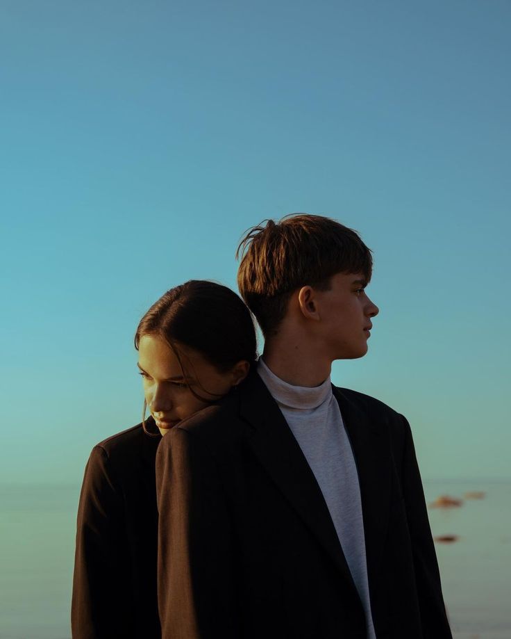 two people standing next to each other in front of the ocean and one person is wearing a black blazer