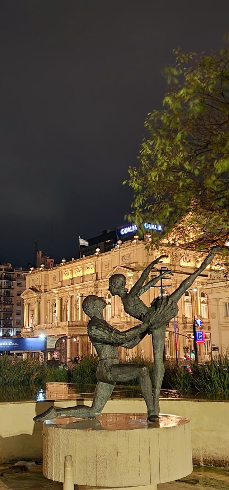 there is a statue in the middle of a park at night with buildings in the background