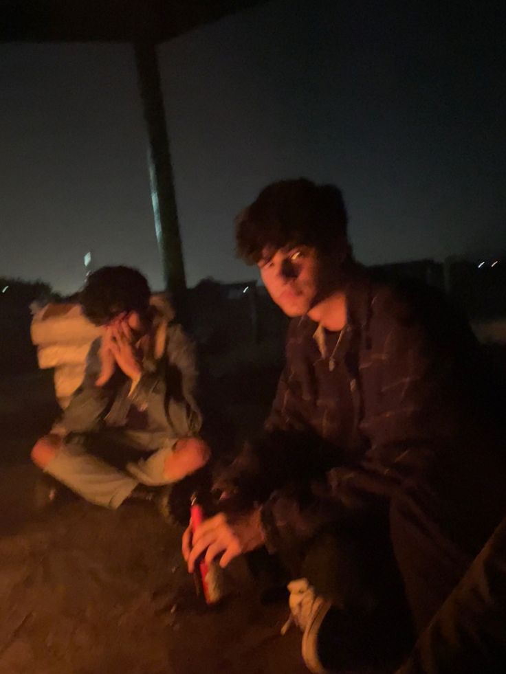 two young men sitting next to each other in front of a campfire at night