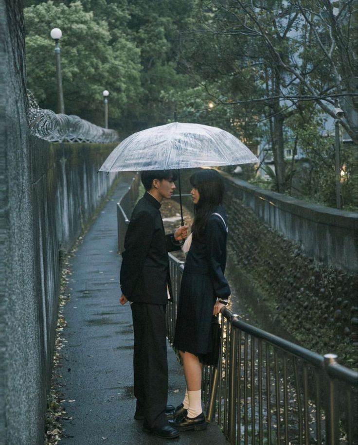 a man and woman standing under an umbrella in the rain on a sidewalk next to a fence