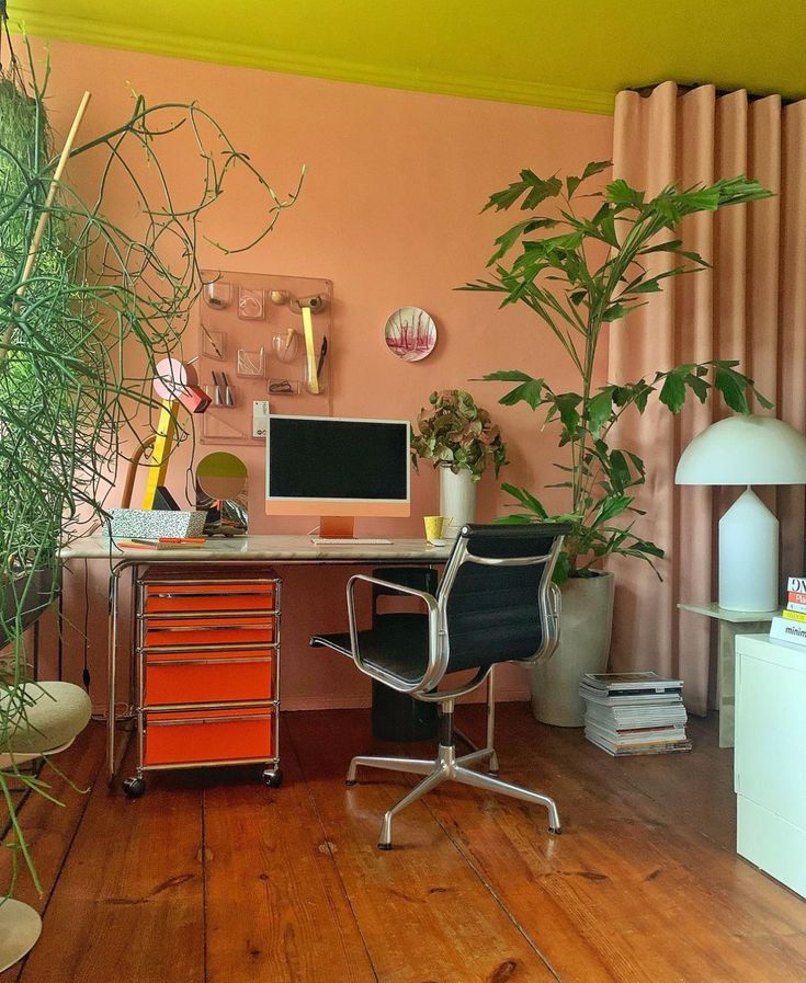 a home office with plants and a computer on the desk in front of a pink wall