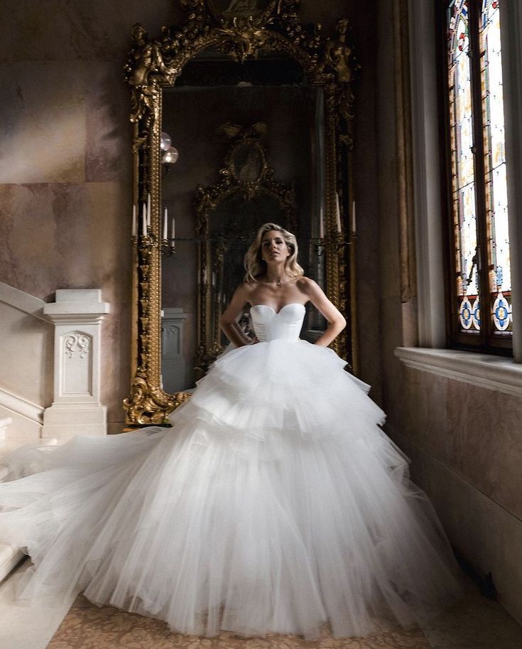 a woman in a white wedding dress standing next to a large mirror and gold framed mirror