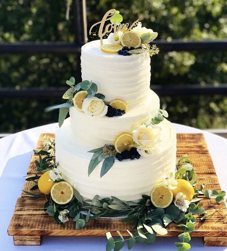 a three tiered cake with lemons and greenery on the top is sitting on a wooden cutting board