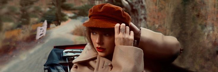 a woman wearing a brown hat and holding her hand up to her face while standing next to a red car