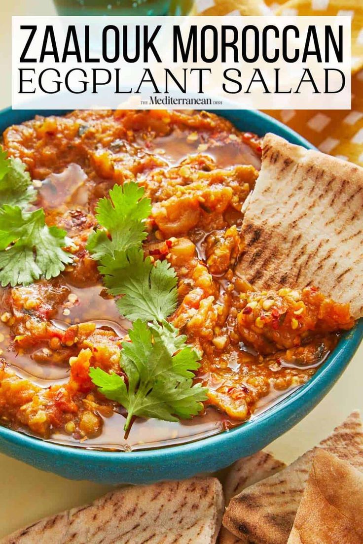 a blue bowl filled with eggplant salad next to pita chips and cilantro