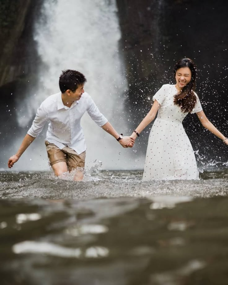 a man and woman holding hands in front of a waterfall with water splashing on them