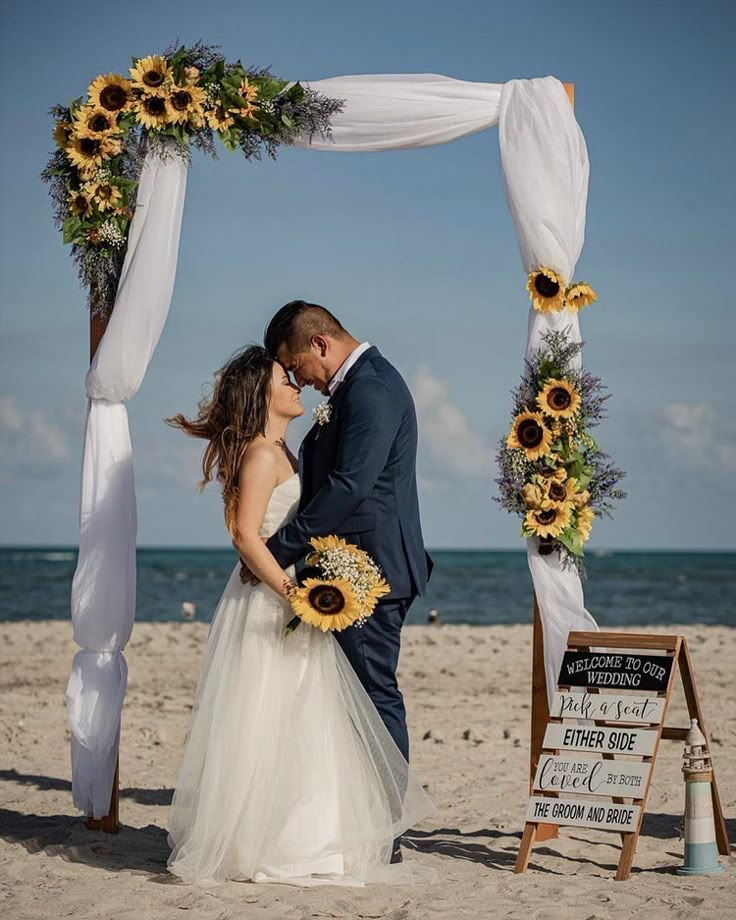 a bride and groom kissing on the beach under an arch decorated with sunflowers