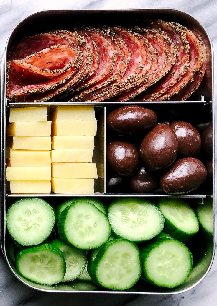 an open lunch box filled with meat, cheese, cucumbers and olives