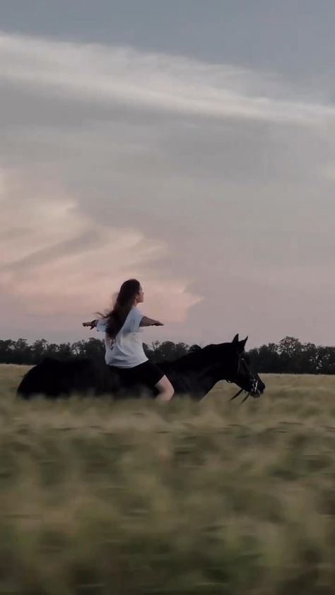 a woman riding on the back of a black horse through a grass covered field at sunset