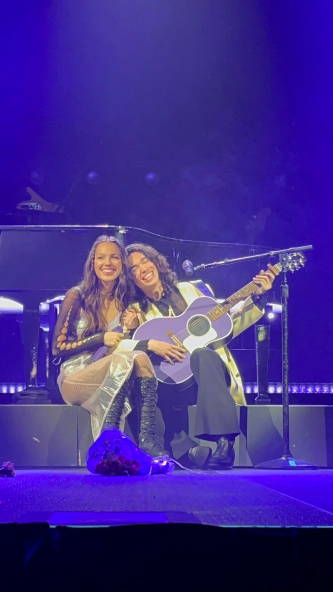 two women sitting on stage with guitars in front of purple lights and one woman is holding the guitar