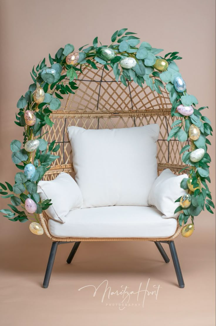 a wicker chair with white pillows and green leaves on it, sitting in front of a pink wall