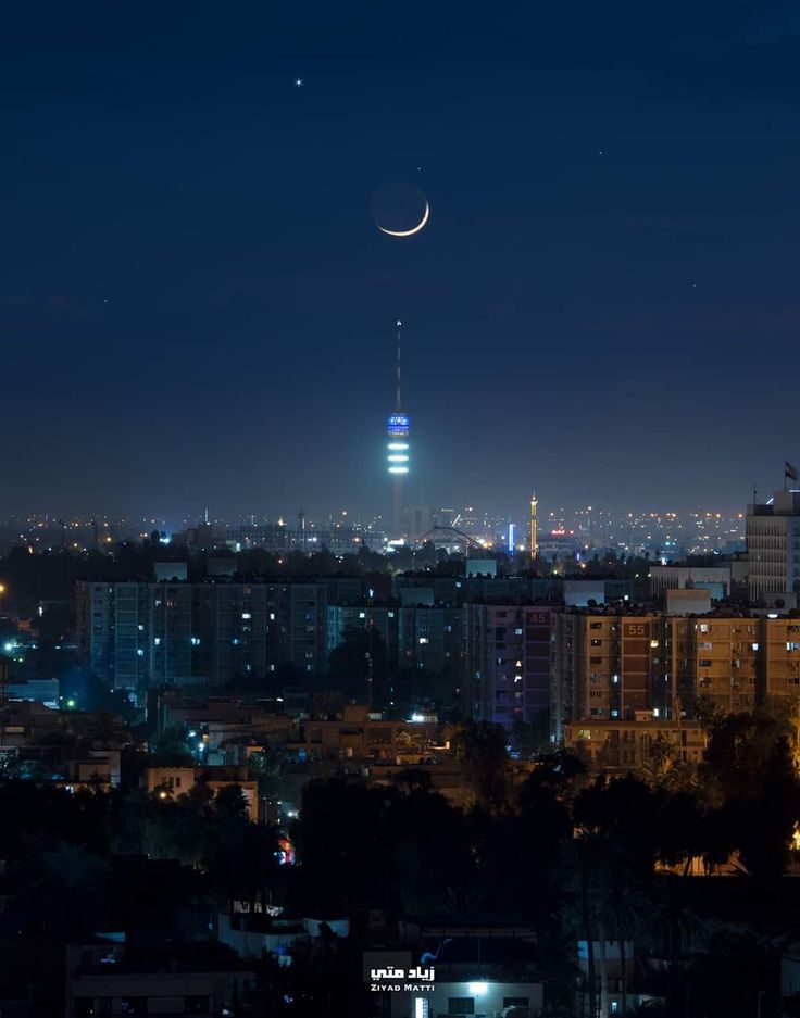 the moon is seen over a city at night with buildings lit up in the background