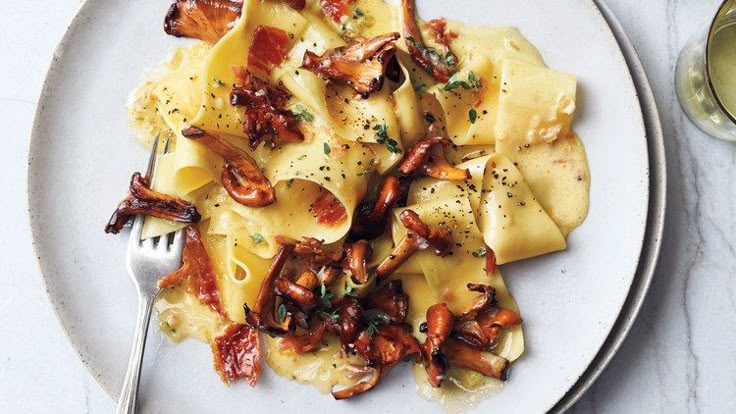 a white plate topped with pasta covered in sauce and toppings next to a glass of wine