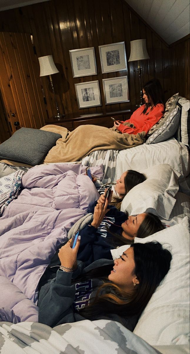 two women laying in bed looking at their cell phones while another woman lays on her side