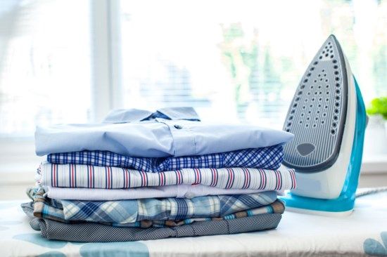 stack of folded clothes next to an iron on top of a table in front of a window