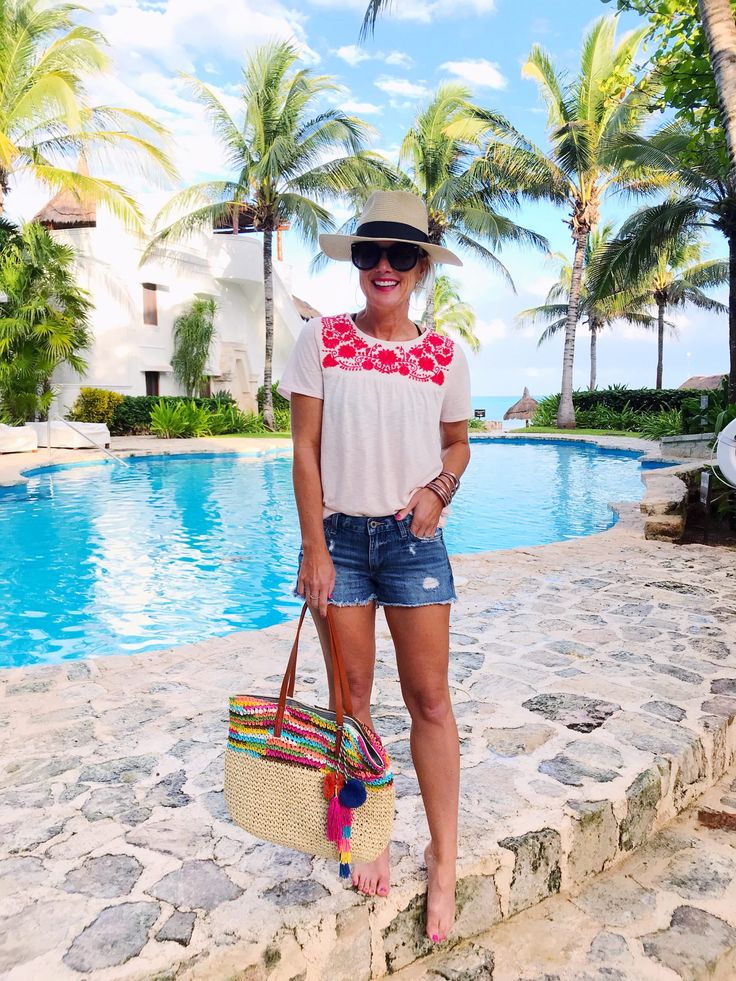 a woman standing next to a swimming pool holding a straw bag and wearing sunglasses with palm trees in the background