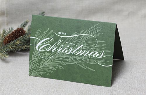 a green christmas card sitting on top of a table next to a pine tree branch