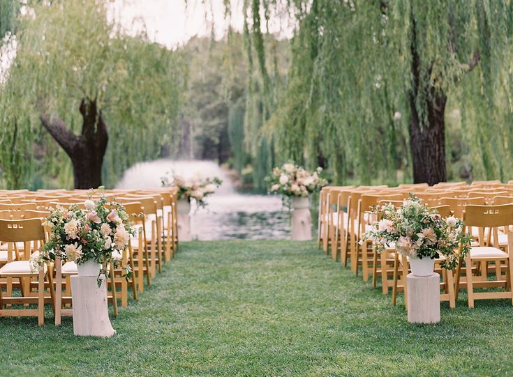 an outdoor ceremony setup with wooden chairs and floral centerpieces on the grass near willow trees