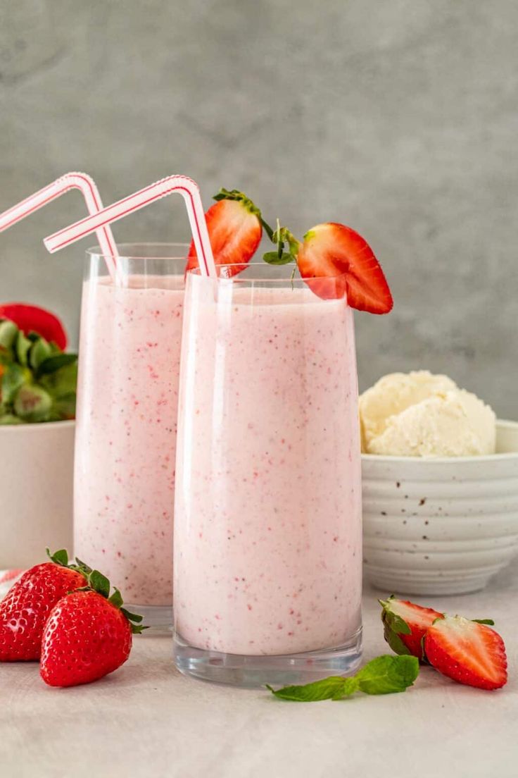 two glasses filled with strawberry smoothie next to bowls of ice cream and strawberries