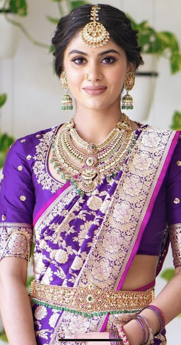 a woman in a purple and gold sari with jewelry on her neck, standing next to a plant