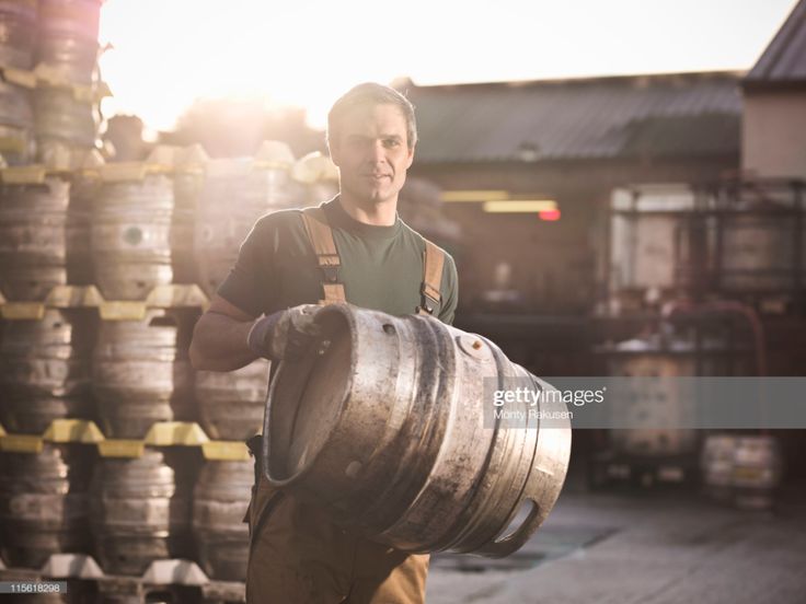 a man carrying a large metal barrel in front of stacks of barrels on the ground