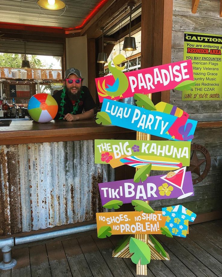 a man standing behind a bar with lots of signs in front of him that read paradise luau party the big kahuna tiki bar