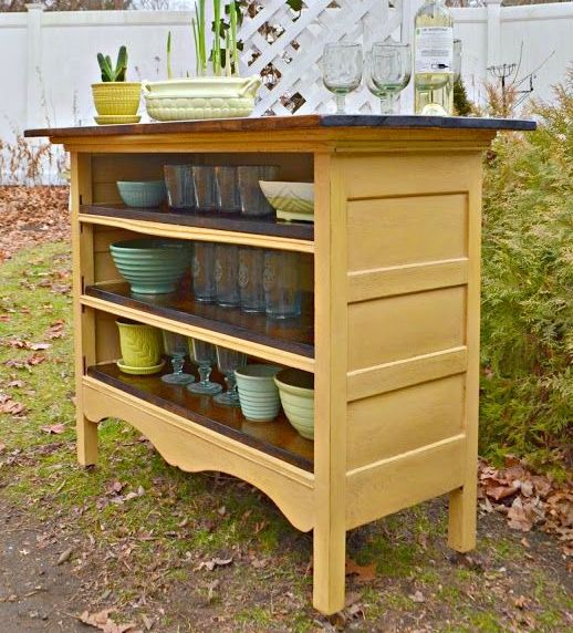 an outdoor kitchen island with pots and bowls on it's shelf, in front of a white picket fence