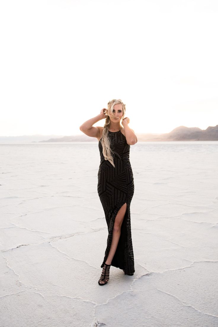 a woman in a black dress is standing on the salt flat with her hands behind her head