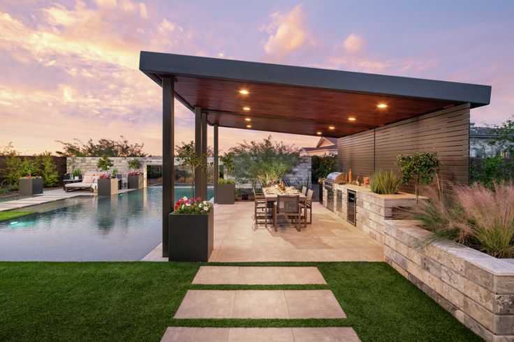 an outdoor kitchen and dining area next to a swimming pool at sunset or dawn with clouds in the sky