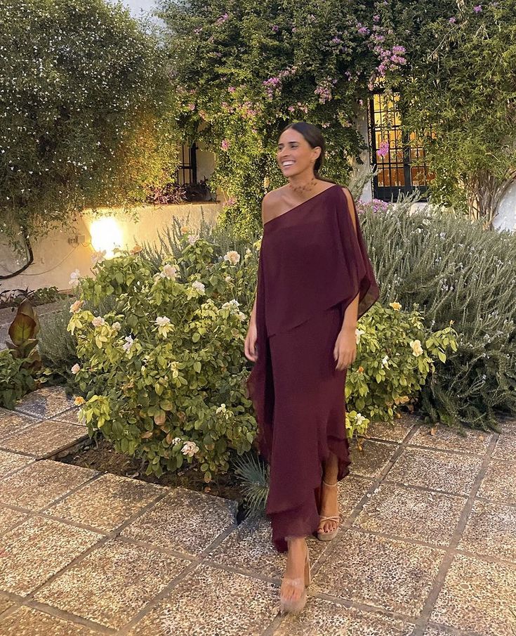 a woman standing in front of some bushes wearing a purple one shoulder dress and tan sandals