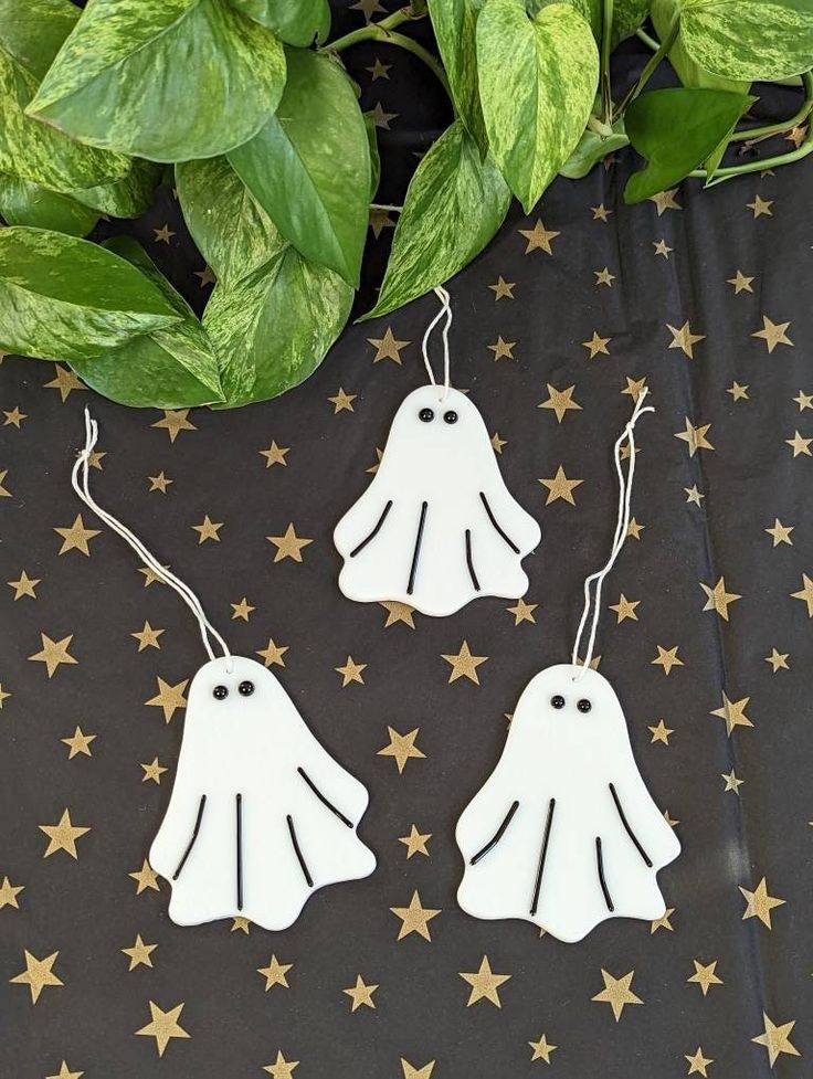 two white ghost shaped earrings hanging from strings on a black background with gold stars and green leaves