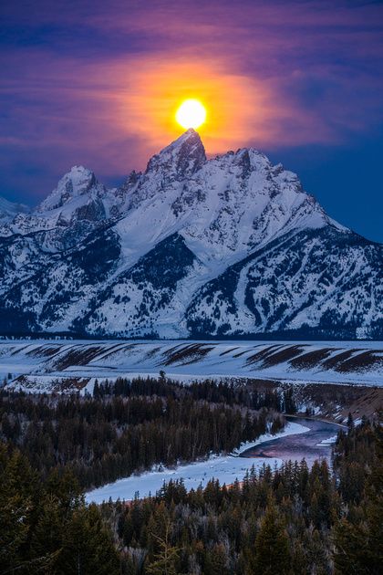 the sun is setting in front of mountains and snow - capped peaks, with pine trees below