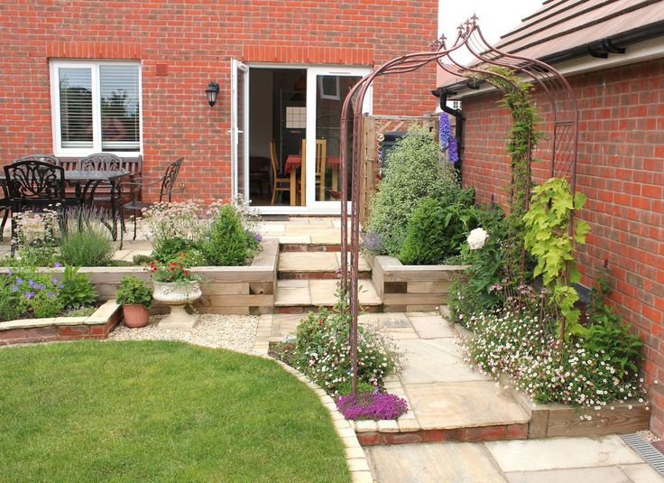 an outdoor patio and garden area with steps leading up to the back door, in front of a red brick house