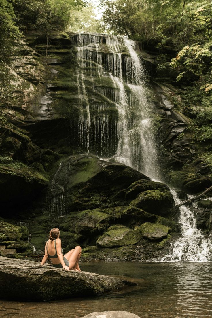 a woman sitting on top of a rock next to a river under a waterfall in the forest