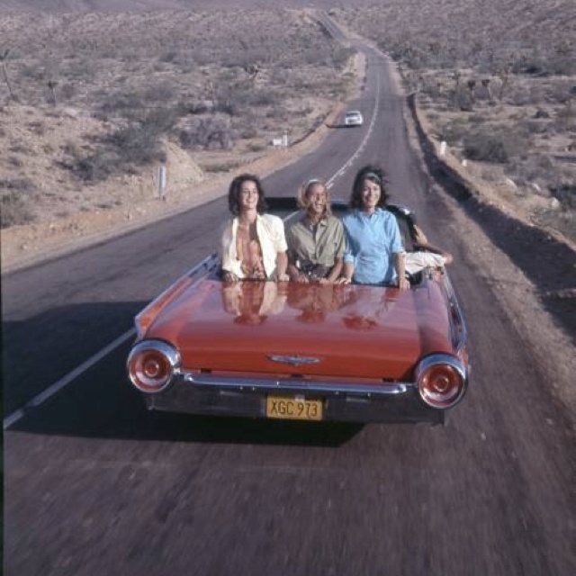 three people are sitting in the back of an old red convertible car on a desert road