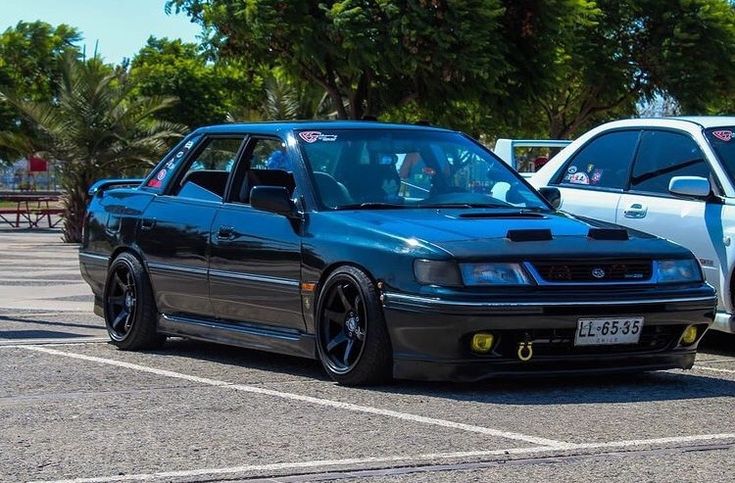 two cars parked next to each other in a parking lot with trees and bushes behind them