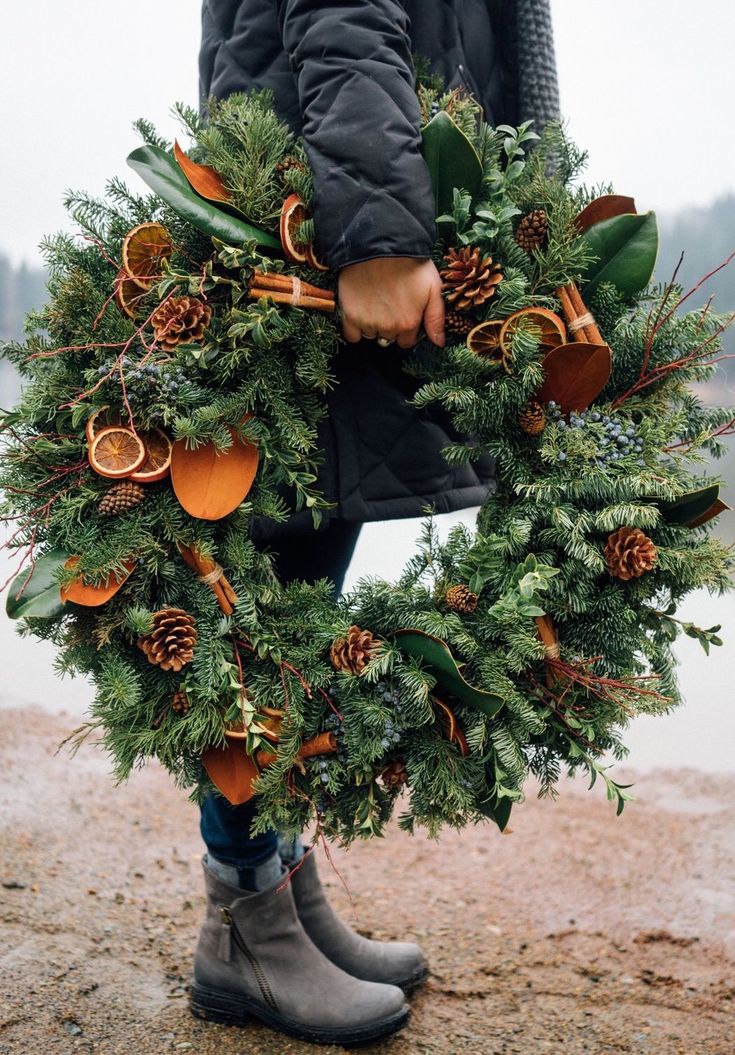 a person holding a wreath with pine cones and oranges on it in front of a body of water