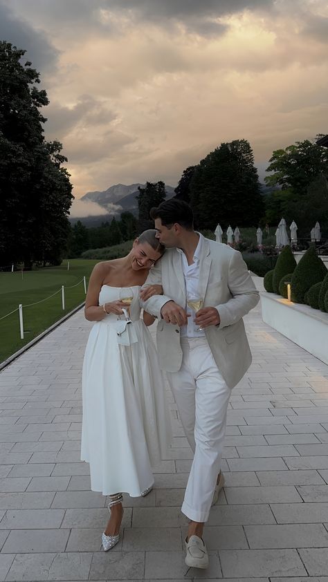 a man and woman dressed in white walking down a walkway with wine glasses in their hands