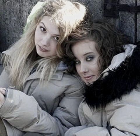 two young women laying on the ground in winter coats, one with her arms around the other's head