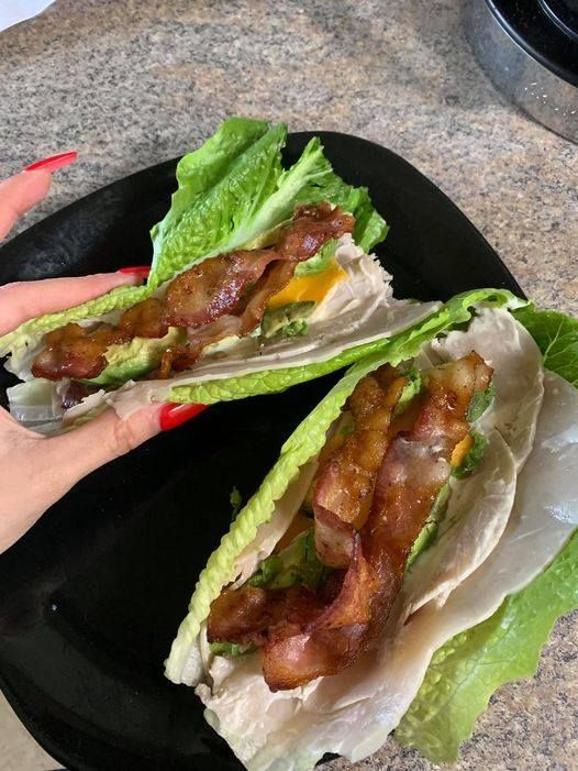 two lettuce wrapped in bacon on a black plate next to a woman's hand