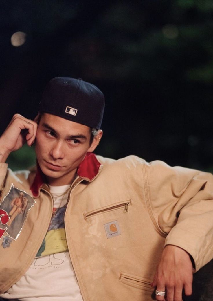 a young man sitting on top of a couch wearing a jacket and hat with his hand to his ear