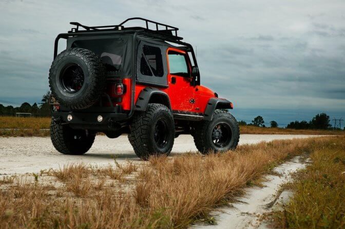 a red jeep driving down a dirt road next to tall grass and dry grass covered ground