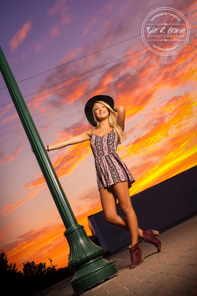 a woman in a dress and hat posing next to a lamp post at sunset with the sun setting behind her