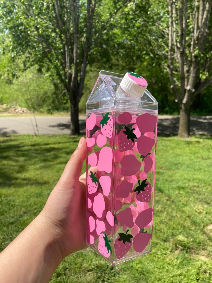 a hand holding up a pink water bottle with strawberries on it and green grass in the background