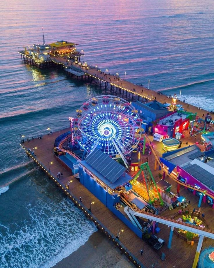 an aerial view of a carnival on the beach at dusk with ferris wheel and rides