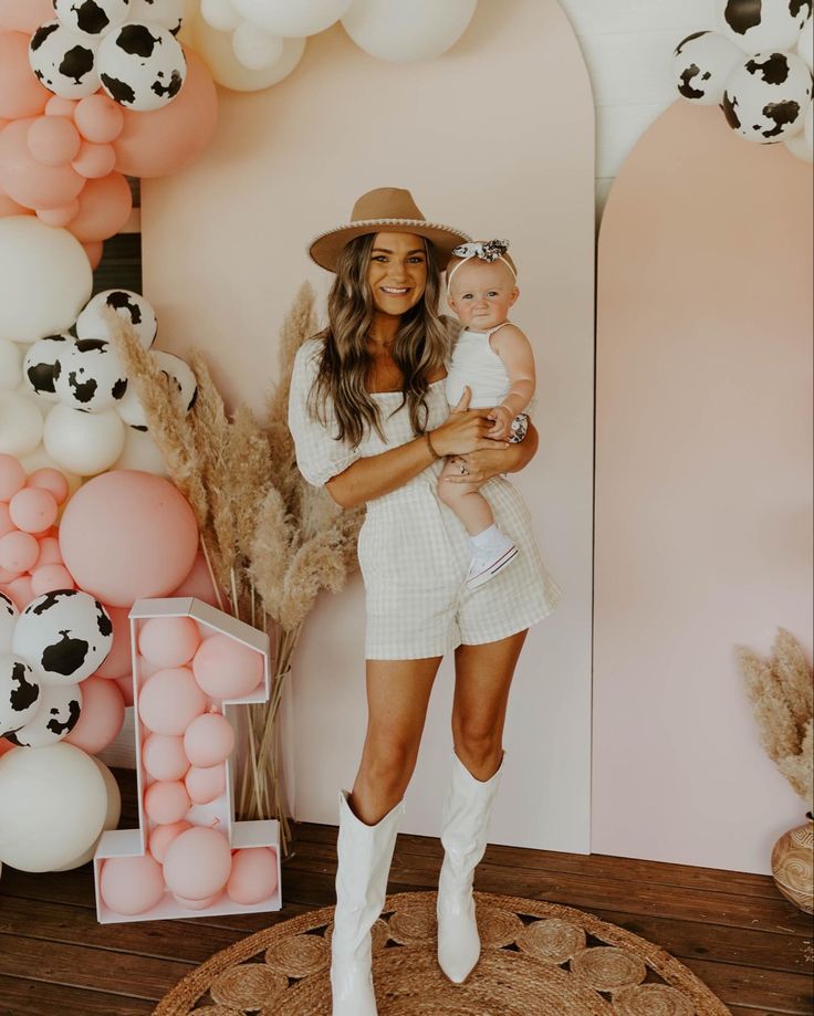 a woman holding a baby standing in front of a wall with pink and white balloons