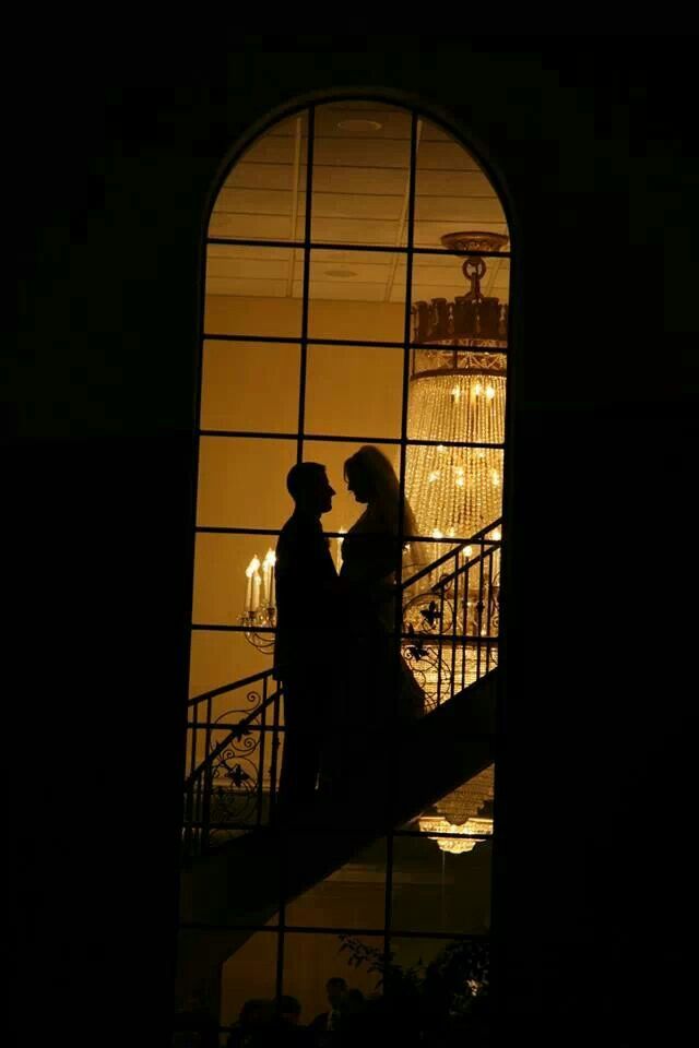 two people standing in front of a window at night with chandelier behind them