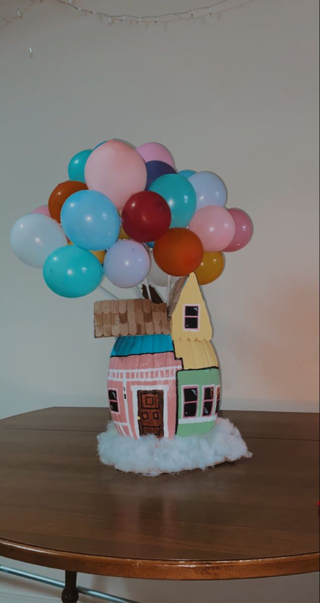 a small house with balloons in the air on top of a wooden table next to a white wall