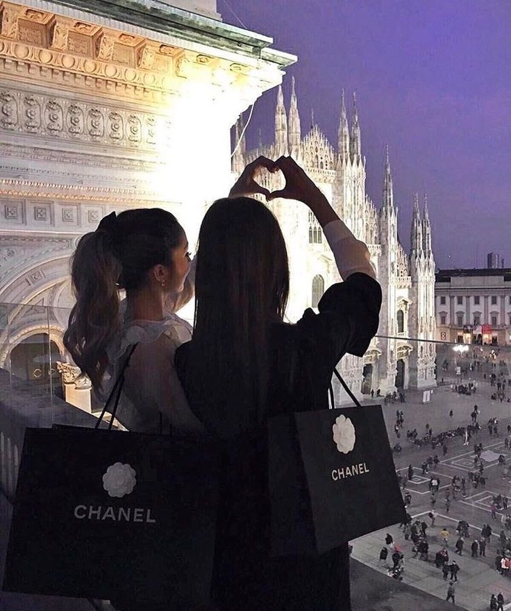 two women standing in front of a large building holding shopping bags and making a heart shape with their hands