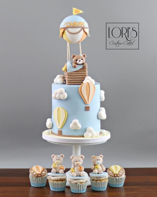 a blue cake with teddy bears and hot air balloons on top is surrounded by cupcakes